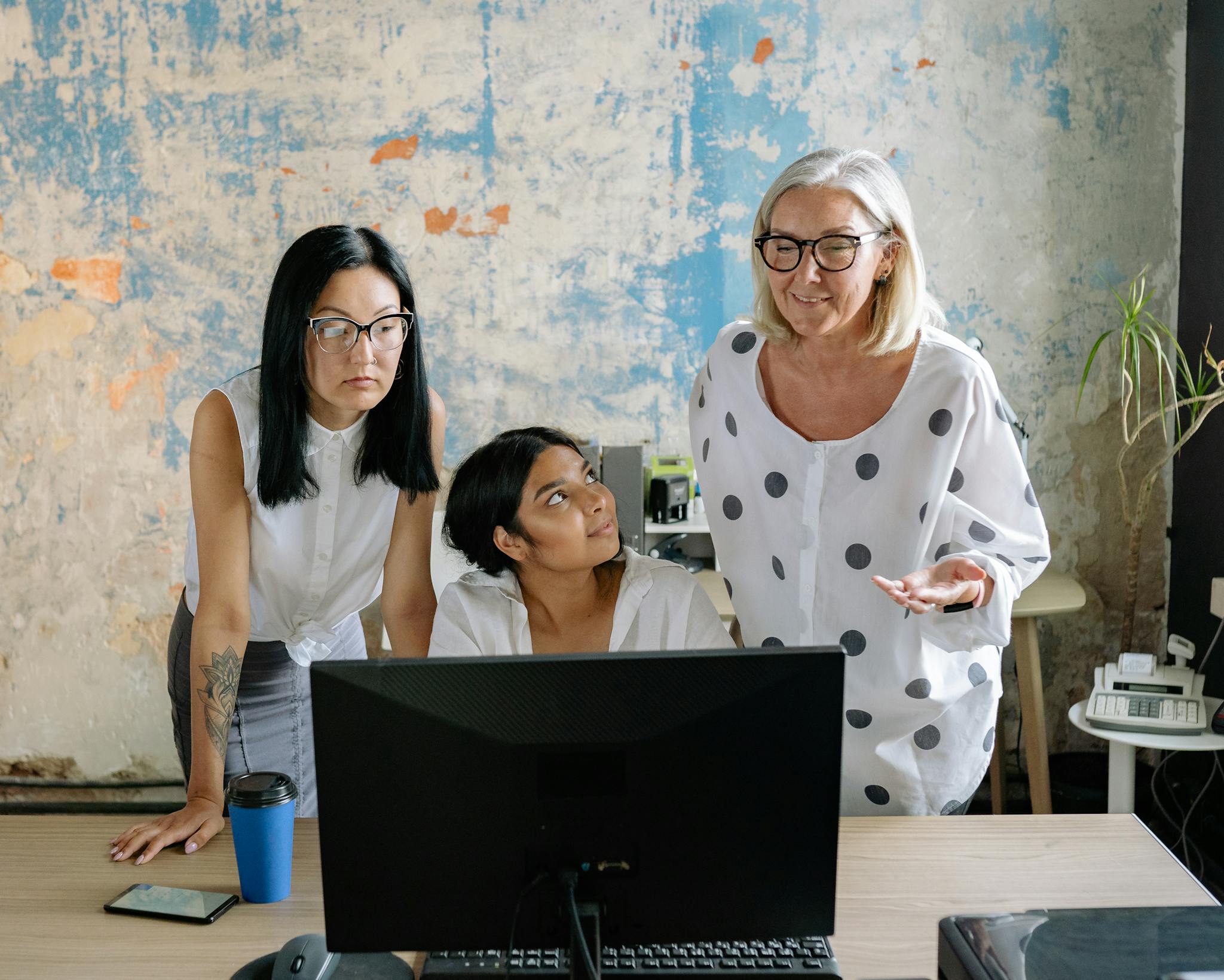 Three diverse businesswomen collaborating at a desk in a modern office environment, focused on a computer screen.